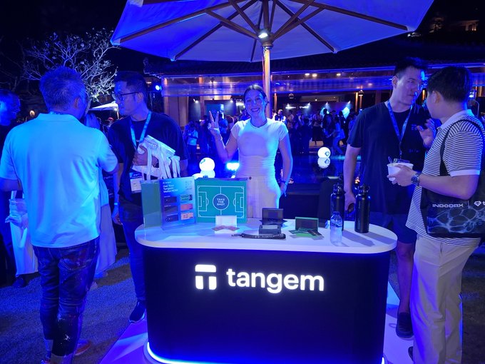 Tangem showcases its crypto wallet brand at Coinfest Asia with an illuminated booth featuring company merchandise and branded giveaways, while team members engage with attendees under ambient blue lighting. The vibrant event atmosphere, with a lively crowd mingling beneath large umbrellas, reflects the "amazing vibes" highlighted in the post.