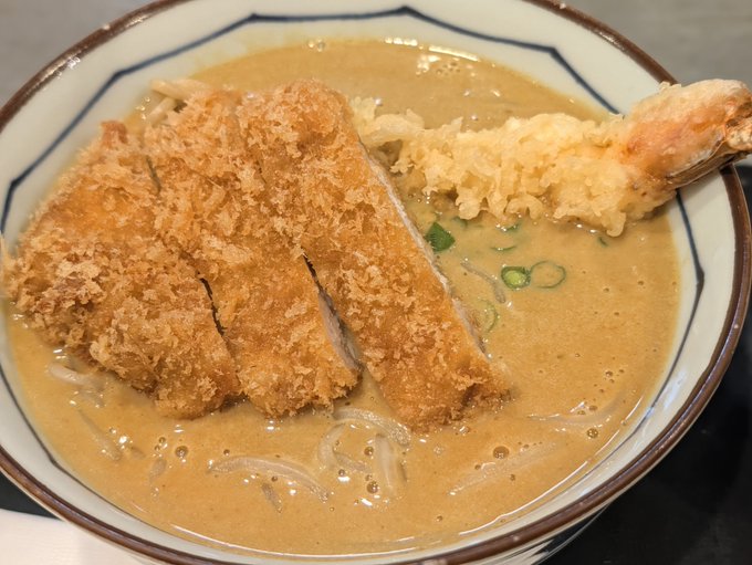 A bowl of curry soba topped with crispy breaded katsu and shrimp tempura, garnished with green onions. The accompanying post celebrates the hearty Japanese meal as a metaphor for perseverance amid the CLARITY Act and crypto market uncertainty, comparing the dish's satisfying "win" to potential bipartisan progress on asset regulation.