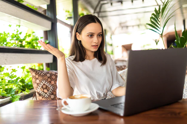 A woman sitting at a wooden table with a laptop and a cappuccino, gesturing with her hand as if speaking during a video call in a bright, plant-filled café setting.