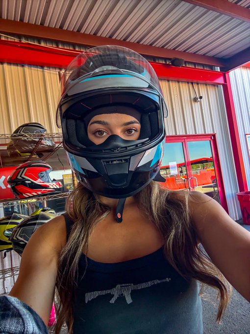 A woman wearing a black tank top takes a selfie while wearing a black, white, and blue motorcycle helmet with the visor raised, standing outside a store displaying helmets.