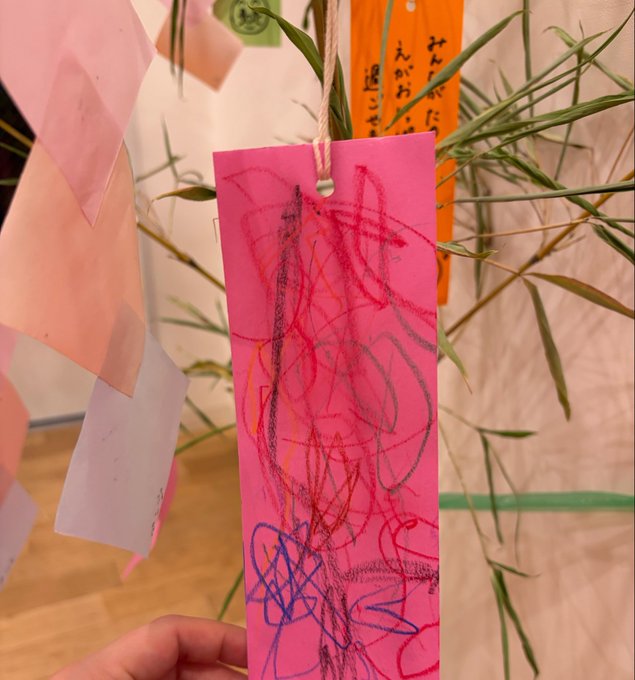 A child's colorful crayon scribble drawing on a pink paper tag hanging from bamboo branches, which appear to be tanabata decorations.