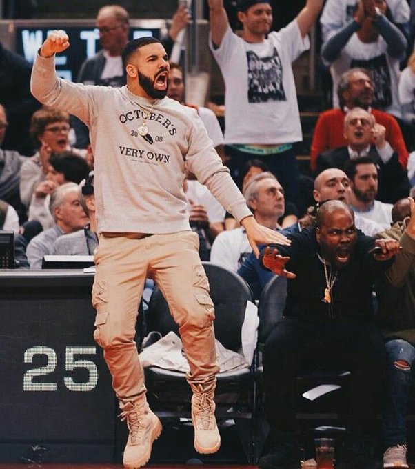 A man wearing a beige sweater reading 'OCTOBER'S VERY OWN' jumping excitedly in the air at what appears to be a basketball game, with fans cheering behind him.