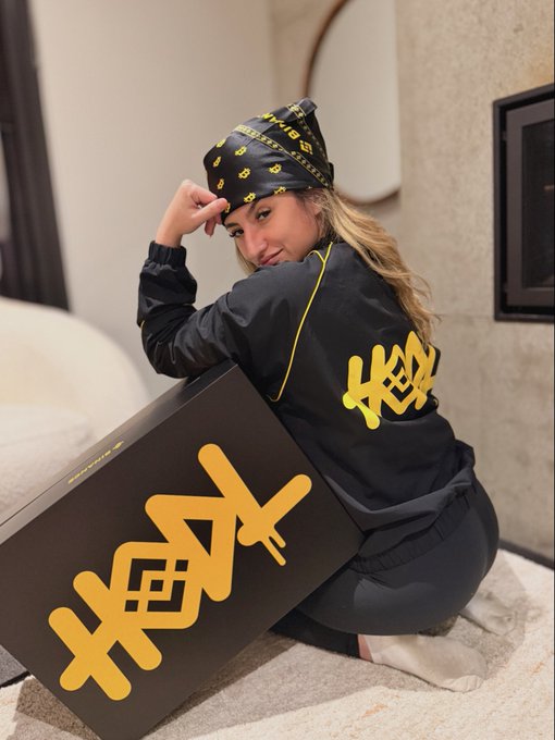 A woman poses with a black and gold Binance-branded box, hinting at an upcoming unboxing video. The bandana and jacket she wears also feature Binance and Bitcoin logos.
