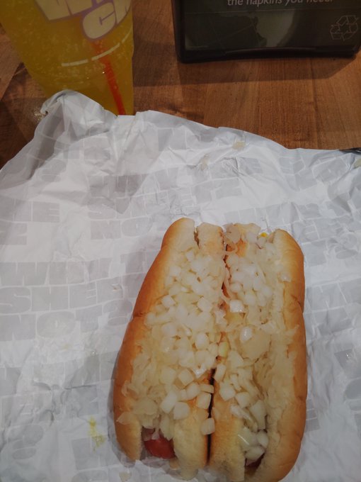A hot dog topped generously with diced white onions sits in a bun on a Subway wrapper, accompanied by an orange beverage in a Subway cup.