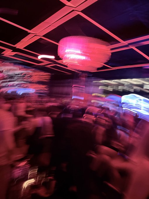 A motion-blurred, abstract photograph of a crowded nightclub interior bathed in vibrant pink and red lighting, featuring a large glowing chandelier.