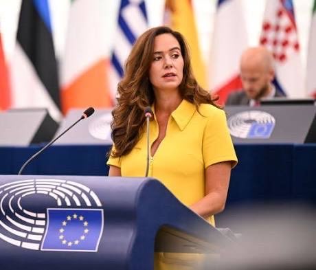 A woman in a yellow dress speaks at a European Parliament podium with the EU flag visible in the foreground and member state flags behind her.