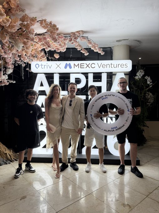 Five people pose together indoors in front of a sign reading 'triv X MEXC Ventures' and 'ALPHA ARENA 2026', with two of them holding a white life preserver labeled 'ALPHA ARENA'.