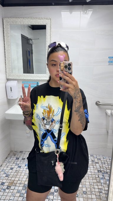 A mirror selfie of a woman in a bathroom wearing an oversized black t-shirt featuring a Vegeta graphic, black biker shorts, and sunglasses perched on her head, holding up a peace sign while taking the photo with a smartphone.
