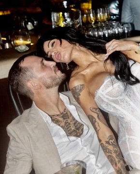 A photograph showing a couple in an intimate moment; a man with heavily tattooed arms and an open white shirt is being kissed by a woman with long dark hair wearing a white lace garment.