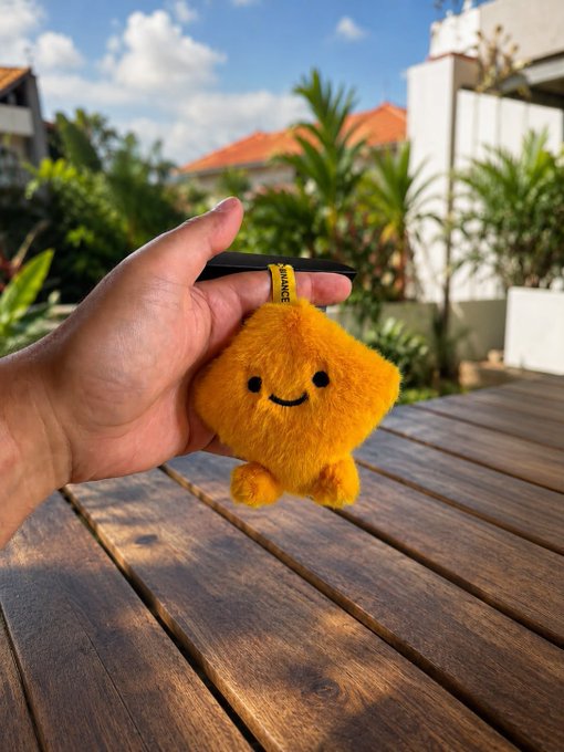 A hand holds up a small, fuzzy yellow star-shaped plush toy with a smiling face, dangling from a black strap labeled "BINANCE," against a sunny outdoor backdrop with a wooden table and tropical greenery. The Binance-branded charm reflects the "Keep Building" weekend message, suggesting enthusiasm and positivity within the crypto/Binance community.