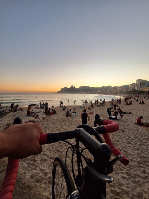 A first-person view from a bicycle with red handlebars on a sandy beach at sunset, with crowds of people and mountains visible in the distance, possibly Ipanema Beach in Rio de Janeiro.
