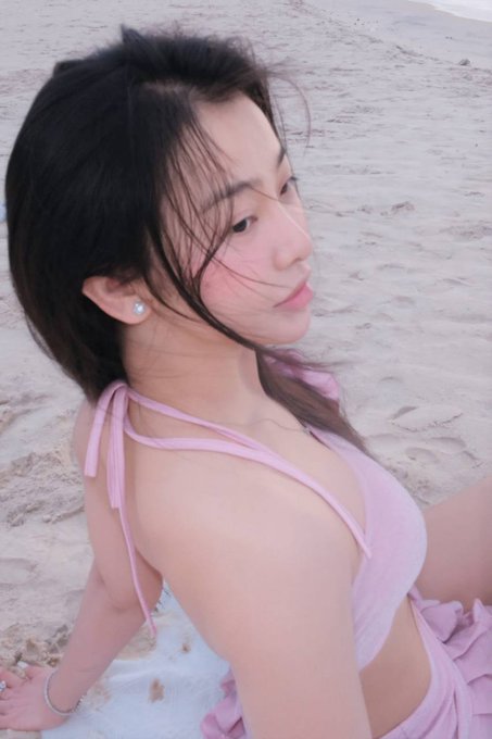 A young woman with dark hair wearing a pink bikini top poses on a sandy beach, looking over her shoulder at the camera.