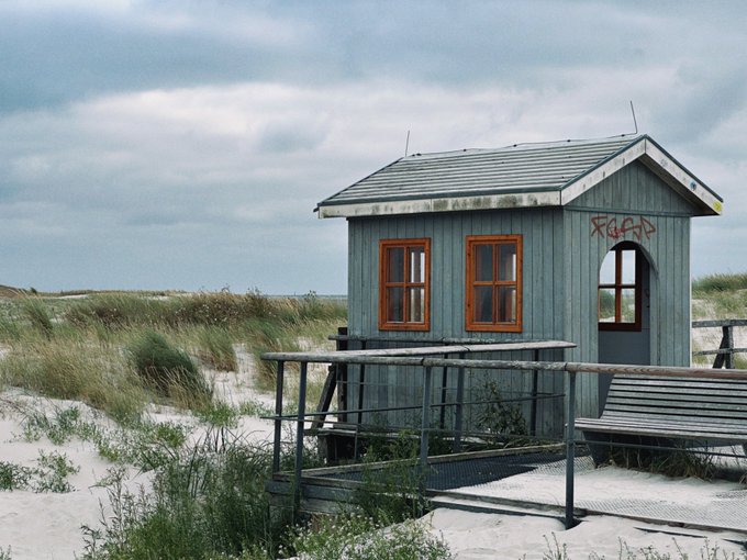 A small blue wooden shack with orange-framed windows and a graffiti tag stands on a sandy dune with grass under a cloudy sky.