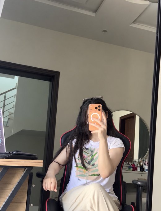 A mirror selfie of a person sitting in a red and black gaming chair, wearing a white t-shirt with a tropical plant graphic, holding an orange smartphone with a magnetic ring attachment.
