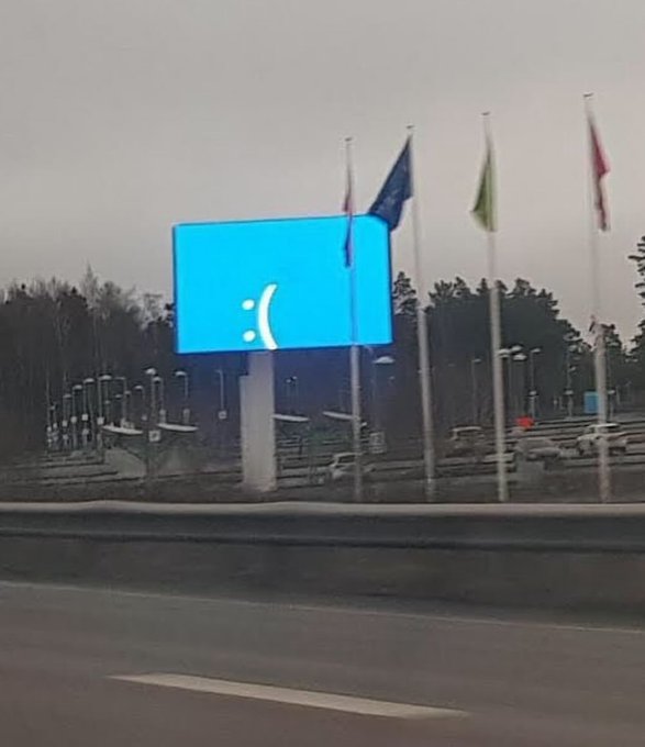 A large outdoor digital billboard displays a sad face emoticon ":(" on a bright blue screen, situated along a road with multiple flags on poles flying in the background under an overcast sky.