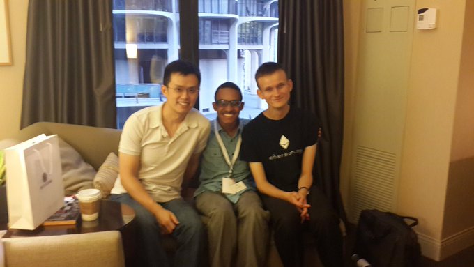 Three young men (Vitalik Buterin flanked by two others) sitting together on a sofa in a hotel room, with an Ethereum-themed white box and a coffee cup on the table beside them.