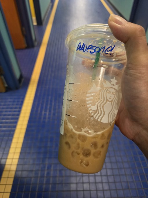 A hand holding a Starbucks iced coffee cup with 'AWESOME!' written on it in blue marker, photographed in a subway corridor.