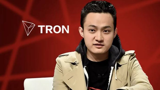 A young man wearing a beige trench coat over a black hoodie is shown with the TRON cryptocurrency logo and wordmark displayed to his left against a red patterned background.