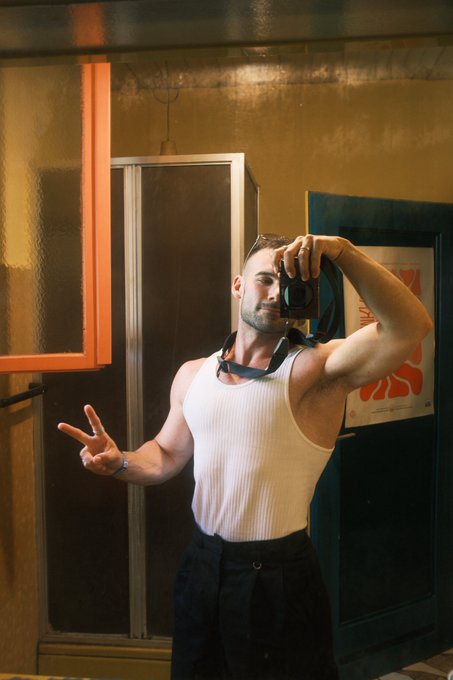 A man wearing a white tank top and black pants takes a mirror selfie with a camera while flashing a peace sign.