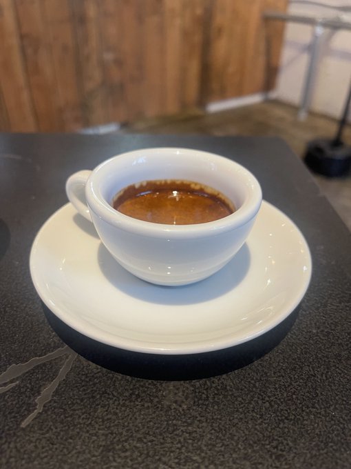 A small white cup of espresso with a rich crema sits on a matching white saucer atop a dark table in a wooden-walled cafe.