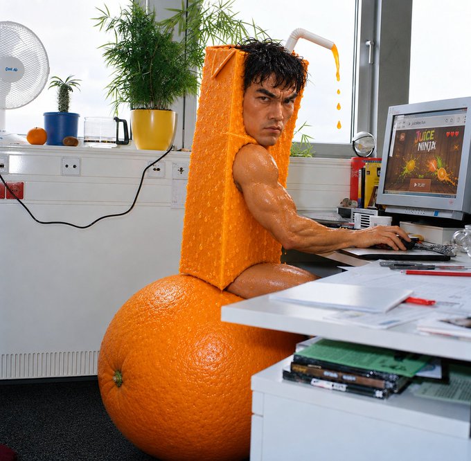 A humorous, staged photo of a man dressed in an orange citrus-themed costume, sitting at a desk using a computer that displays a game called 'JUICE NINJA'.