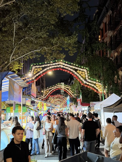A crowded street festival at night, illuminated by arched strings of colorful lights, with vendor stalls lining the walkway.