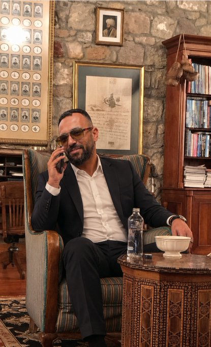 A bearded man in a dark suit and sunglasses sits in an ornate striped armchair within a richly decorated study featuring stone walls, framed artwork, and a wooden bookcase, talking on a phone while a water bottle and bowl rest on a carved wooden table beside him. The Turkish caption "Acele eden değil, hazırlanan kazanır" ("It is not the one who hurries, but the one who prepares who wins") reflects his composed, deliberate demeanor as he calmly conducts business in a refined setting that suggests careful preparation and strategic thinking.