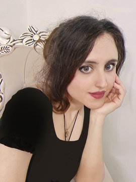 Portrait of a young woman with dark hair, wearing a black top and delicate pendant necklaces, resting her chin on her hand against a decorative silver-toned background.