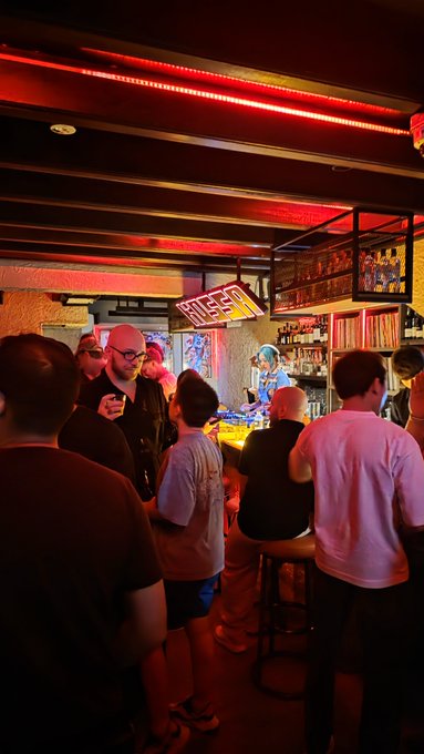 A dimly lit, crowded bar scene with red neon lighting, where patrons are gathered around the counter where a bartender is preparing drinks.