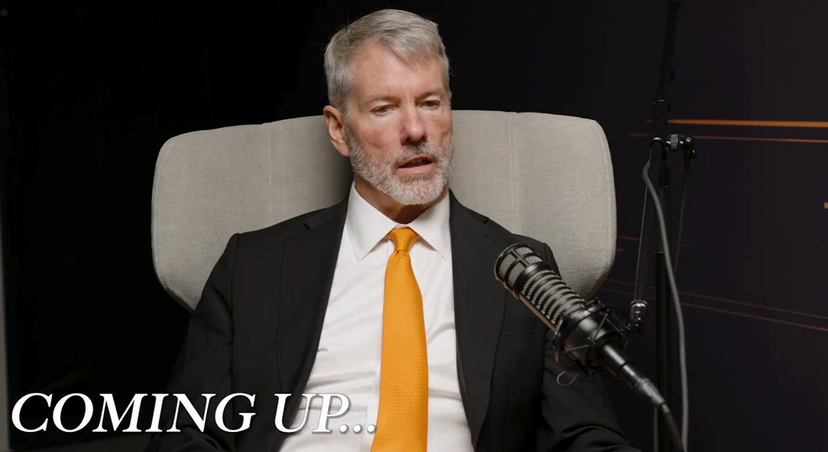 The image features a man wearing a suit and tie sitting behind a microphone, with the text "coming up..." written above him. The focus of the image seems to be on the man's attire and his position as a speaker or presenter at an event related to Bitcoin or cryptocurrency.