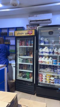 A small shop interior showing refrigerated display cases stocked with various drinks and dairy products, with a person visible working behind the counter.