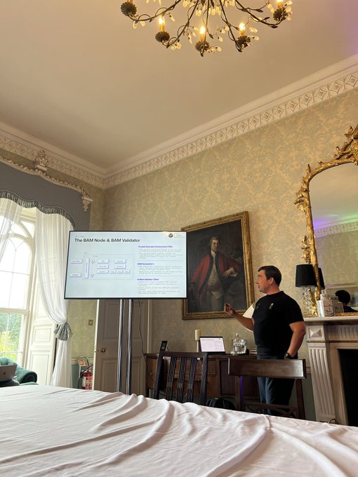 A man in a black t-shirt stands presenting next to a screen displaying 'The BAM Node & BAM Validator' in an ornate room with patterned wallpaper, a portrait painting, a chandelier, and a bed in the foreground.