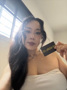 A selfie of a woman with long dark hair wearing a white tube top, holding a black card with gold text reading 'GOLD'.