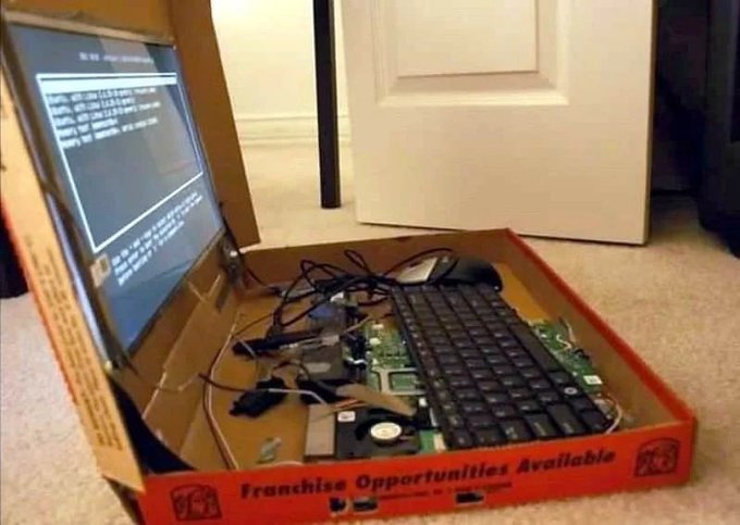 A DIY computer setup with a monitor and keyboard resting on top of exposed motherboard components inside a cardboard box labeled 'Franchise Opportunities Available' on a carpeted floor.