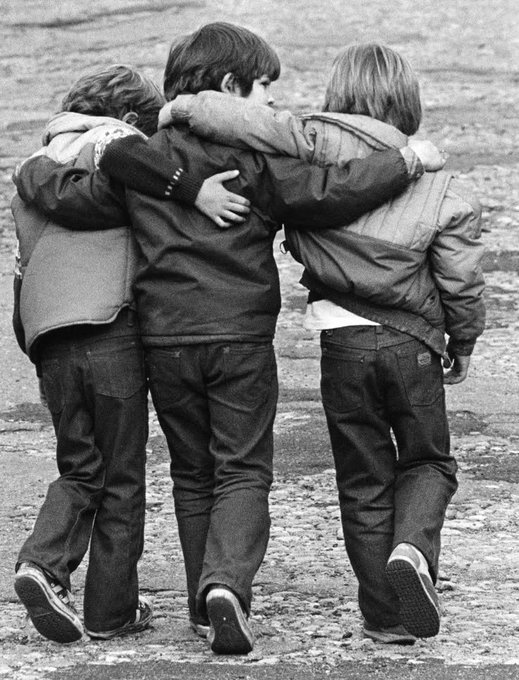 A black-and-white photograph shows three boys walking together with their arms around each other's shoulders, viewed from behind.
