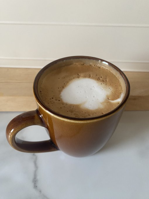 A brown ceramic mug filled with a frothy coffee drink sits on a white marble countertop, with foam art resembling a small cloud or shape on the surface.