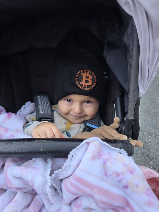 A smiling child sits in a stroller wearing a black beanie with the orange Bitcoin '₿' logo, holding a small stuffed animal.