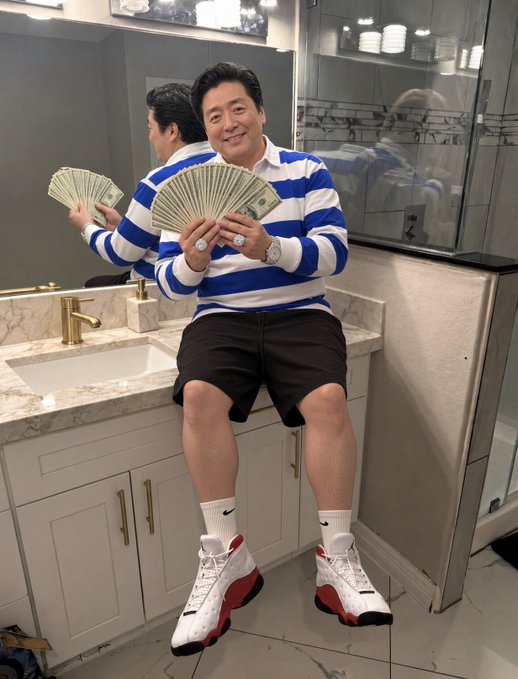 A man wearing a blue and white striped sweater and black shorts sits on a bathroom counter holding a fan of US dollar bills, wearing white Nike socks and red and white sneakers, with a watch and ring visible on his hands.