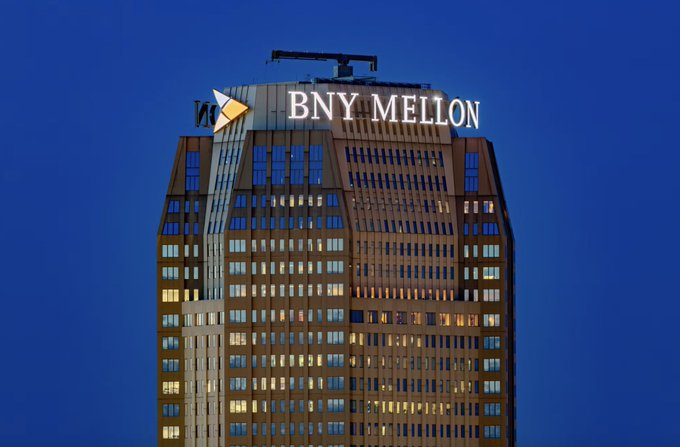 The top of a tall building at dusk with illuminated 'BNY MELLON' signage against a dark blue sky.