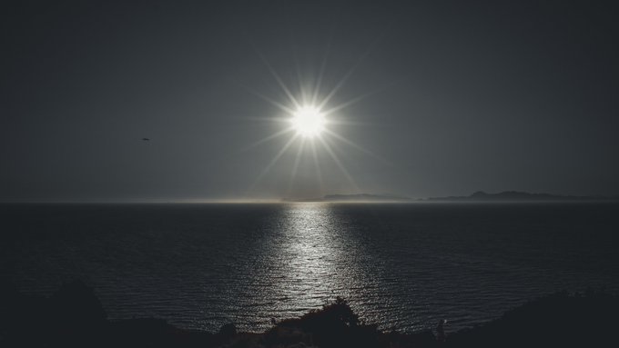 A stunning sunset or sunrise over a dark ocean, with the sun creating a starburst effect and reflecting a bright path across the water toward a distant landmass silhouette.