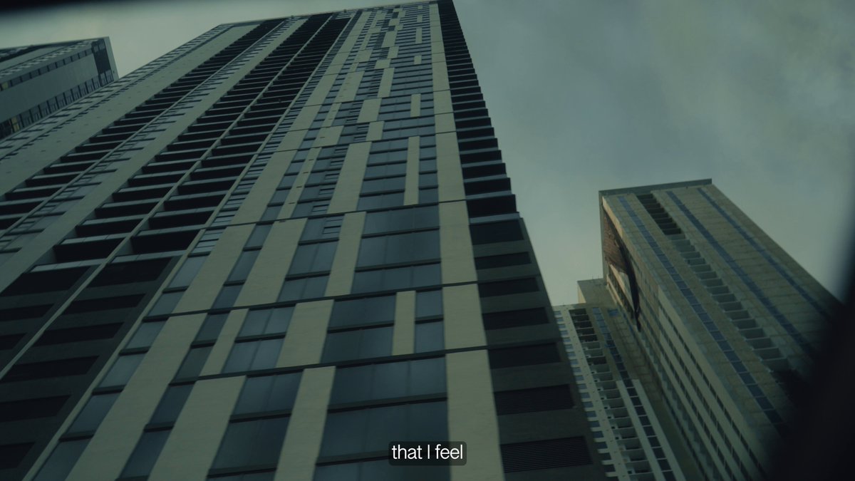 The image shows a tall building with many windows, giving an impression of a modern architectural design. Below this skyscraper, there are other buildings that appear smaller due to their height. The sky above them has some clouds, adding depth to the scene. In the bottom right corner of the image, there is text that reads "Building Sushi in Public - Episode 9 out now on YouTube". This suggests that a new series or movie about sushi-making might be released soon, and it could potentially feature this building as one of its filming locations.
