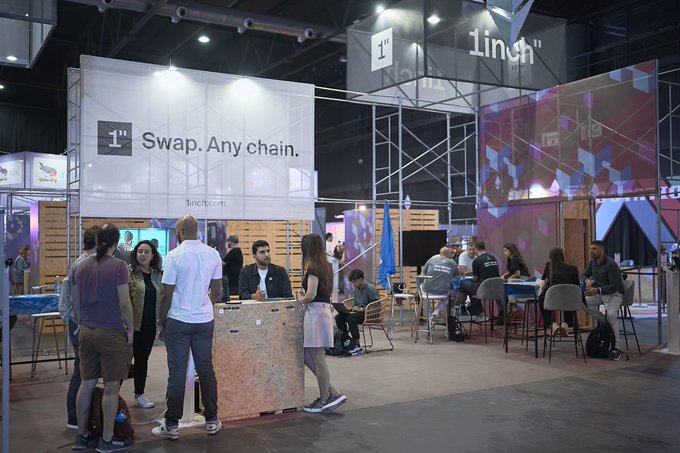 The image features a large room filled with people gathered around tables, engaging in conversations and networking. Some of them are standing while others are sitting at chairs. The floor is made of tiles, adding to the formal atmosphere of the event. A sign prominently displayed within the space reads "1 T-minus 3 days to @ethconf" which indicates that an electronic conference related to Bitcoin will take place soon. Several people in the room appear to be carrying backpacks and laptops, suggesting they may be attendees or vendors at this gathering. The overall vibe of the image appears to be focused on business, networking, and possibly discussing emerging trends like cryptocurrency.