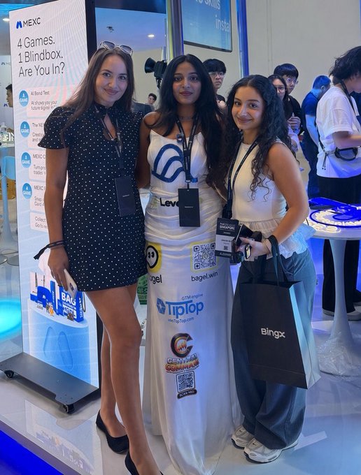 Three young women pose at what appears to be a crypto/Web3 conference (MEXC booth with text '4 Games, 1 Blindbox, Are You In?'), wearing promotional outfits and lanyards including 'TiPlop' and holding a 'BingX' bag.