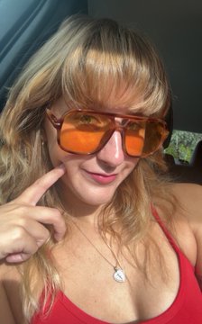 A woman with blonde hair and bangs wears orange-tinted aviator sunglasses and a red top, posing while pointing at her face.