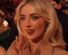 A young blonde woman with glamorous makeup and glossy lips claps her hands at what appears to be a formal event.