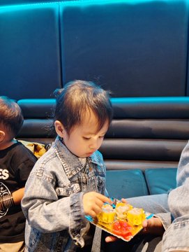 The image shows a young child wearing a denim jacket, holding a plate with sushi on it. The background features a blue wall that complements the color of the child's jacket. There are two other people present in the scene, one standing behind the child and another person partially visible at the top right corner. The post is about Bitcoin and Web3, which seems to be related to the sushi or food served by the restaurant.
