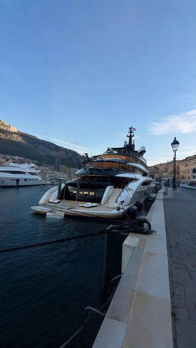 A large luxury yacht docked at a harbor with mountains in the background and a clear blue sky, viewed from a stone pier with an old-fashioned lamppost.
