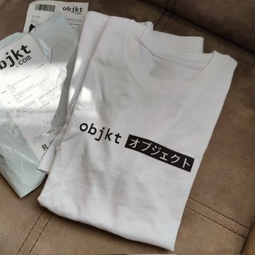 A folded white t-shirt with the text 'objkt' and Japanese characters 'オブジェク' (object) printed in a black box, sitting next to its shipping packaging from objkt.com on a brown couch.