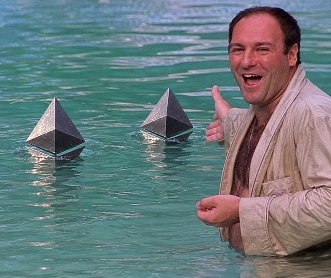 Actor Kevin Spacey in a tan bathrobe laughs while in a swimming pool next to two pyramid-shaped objects floating in the water.