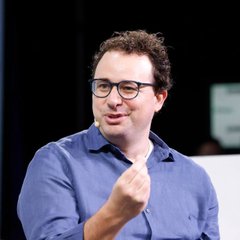 A man with curly hair and glasses, wearing a blue button-down shirt, is speaking while gesturing with one hand.